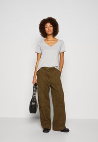 Gray v-neck t-shirt paired with loose olive green cargo pants. Model holds a black bag with silver accents, standing on a light gray background.