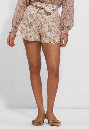 Vrouw die een bloemenprint korte broek met hoge taille en een strikceintuur draagt, samen met beige schoenen met hak en grote strikken, staand tegen een effen achtergrond.