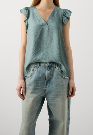 Vrouw die een lichtblauw mouwloos blouse met V-hals draagt, met gerimpelde schouders en vervaagde blauwe jeans met hoge taille.