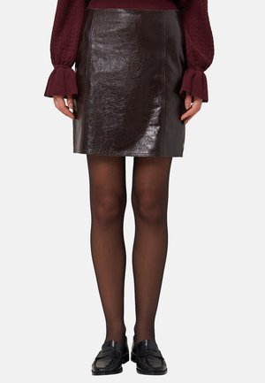 Jupe mini en cuir marron avec une finition brillante et des coutures plates, associée à des collants noirs et des mocassins, sur un fond uni.
