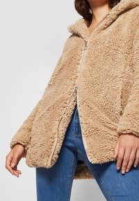 Kvinde iført en beige, fluffy zip-up jakke og blå jeans, set fra brystet til midten af låret, med hænderne afslappet ned langs siden.