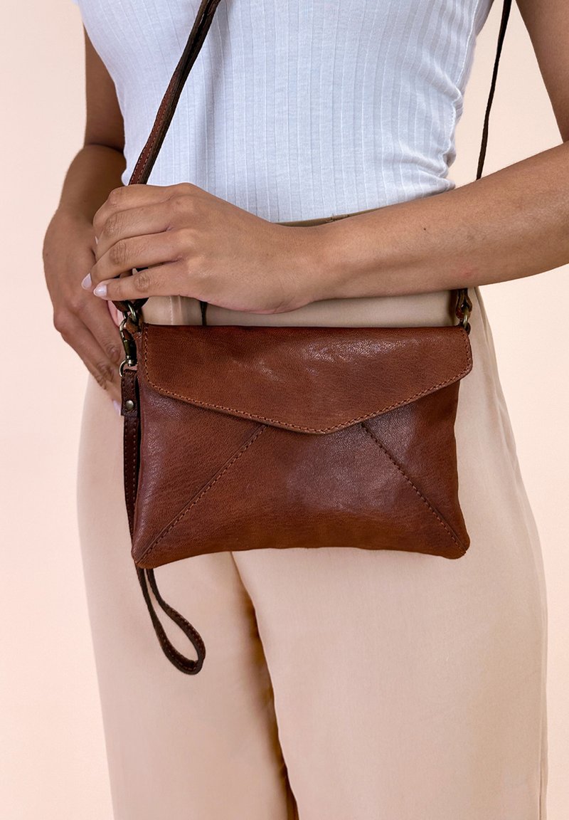 Borsa a tracolla in pelle marrone con flap triangolare, texture liscia e tracolla removibile, tenuta in mano su uno sfondo neutro.