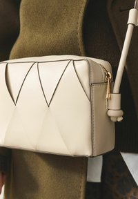 Sac en cuir beige texturé avec un design géométrique, doté d'une fermeture éclair dorée et d'un accent de bandoulière arrondi. Positionné sur une tenue sombre.