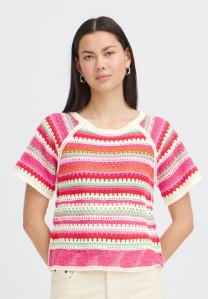 Žena s dlhými tmavými vlasmi, ktorá nosí strieborné kruhové náušnice a farebný pruhovaný háčkovaný top s krátkymi rukávmi na jednoliatom pozadí.