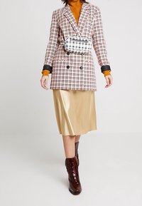 Blazer à carreaux à double boutonnage, associé à une jupe midi en satin doré et des bottes bordeaux montantes jusqu'aux genoux. Tient un petit sac noir et blanc.