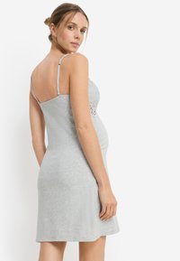 Γκρι φόρεμα εγκυμοσύνης με ρυθμιζόμενες τιράντες, λεπτομέρειες δαντέλας στο πάνω μέρος, εφαρμοστή σιλουέτα και φαρδύ τελείωμα. Λείο ύφασμα από βαμβακερό μείγμα.