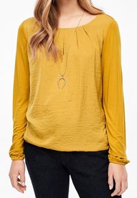 Senapsgul långärmad blus med texturerad finish, rund halsringning, veck vid bröstet och subtila sammanknäppta manchetter.