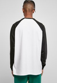 Langärmliges T-Shirt mit Raglanärmel, bestehend aus schwarzen Ärmeln und einem weißen Körper. Hergestellt aus Baumwolle, mit einer glatten Textur und abgerundetem Saum.
