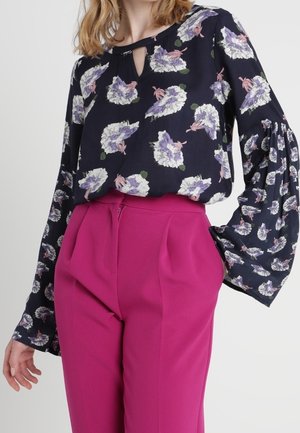 Femme portant un chemisier bleu marine à motifs floraux avec un col en trou de serrure et des manches volumineuses, associé à un pantalon taille haute rose vif sur mesure.
