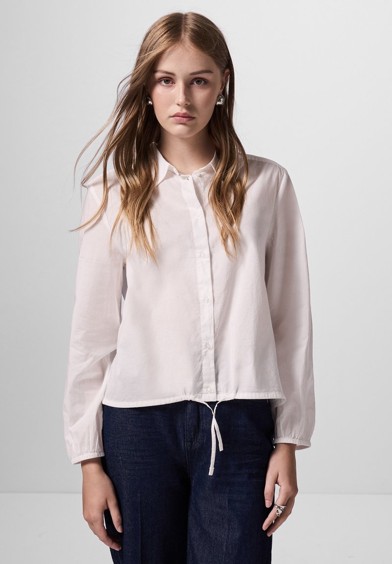 Weiße, langärmelige Hemdbluse aus leichtem Stoff mit einem Kordelzugsaum und klassischem Kragen, kombiniert mit dunklen Jeanshosen.