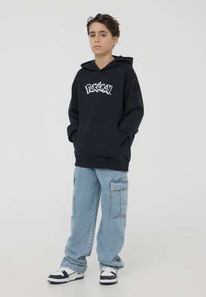 Felpa nera con logo "Pokémon" in bianco, dotata di tasca frontale. Abbinata a jeans cargo blu chiaro e scarpe da ginnastica nere e bianche.