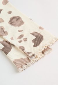 Legging en tissu côtelé crème avec des taches irrégulières imprimées vache marron clair et des ourlets à volants sur fond blanc.