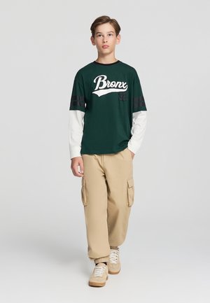 Maglietta verde a maniche corte con testo bianco "Bronx 92", sovrapposta a una maglietta bianca a maniche lunghe. Pantaloni cargo color sabbia con tasche laterali e sneakers beige.