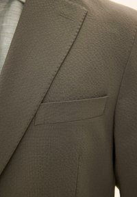 Zbliżenie na fakturowany klapę marynarki w oliwkowo-zielonym kolorze z przeszytym dziurkiem na guzik i kieszenią na piersi z listwą.