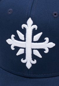 Laevastiku baseballi müts, millel on suur valge tikandiga embleem, millel on keerukad lehe- ja ristikujundid, struktuurne kuju ja sile kangas.