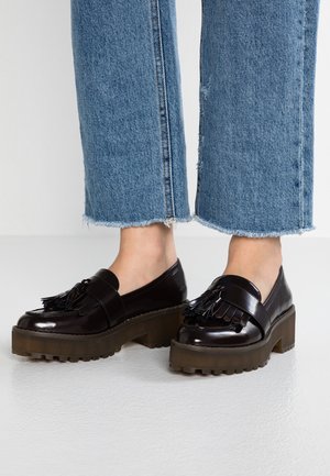 Mocassins noirs en cuir verni avec pampilles et semelles épaisses marron à crampons, portés avec un jean bleu à ourlet effiloché sur fond blanc.