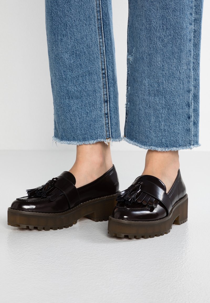 Mocassins noirs en cuir verni avec pampilles et semelles épaisses marron à crampons, portés avec un jean bleu à ourlet effiloché sur fond blanc.