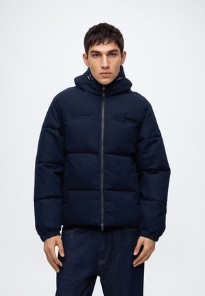 Jeune homme portant une veste matelassée bleu marine avec une capuche, debout devant un fond clair uni, faisant face à la caméra avec les mains détendues.