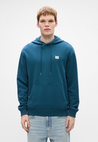 Mėlynas džemperis su kapučinu, virvute, priekyje kišene ir siuvinėtu logotipu. Pagamintas iš minkštos medžiagos, turi laisvą siluetą ir sutraukiamas rankogalius.