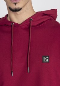 Felpa color bordeaux con cappuccio regolabile. Presenta una toppa quadrata con logo e cordini con punte metalliche. Il tessuto sembra morbido e ha una superficie liscia.