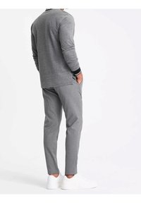 Chemise grise à manches longues avec un poignet noir, associée à un pantalon gris clair fuselé et des baskets blanches, vue de dos.