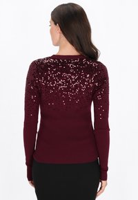 Pull bordeaux à manches longues, présentant un motif de sequins sur les épaules, une texture côtelée sur le corps et un col rond.