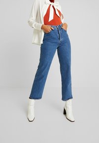 Jeansy o wysokim stanie i prostych nogawkach w średnim niebieskim deninie, zestawione z czerwoną bluzką i białymi botkami na obcasie. Całość dopełnia biała marynarka.