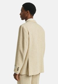 Homme avec des cheveux courts et bouclés portant une veste de costume texturée beige et un pantalon assorti, se tenant de profil contre un fond blanc uni.