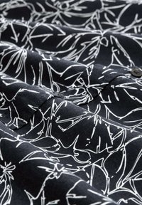 Tissu noir avec un motif de dessin floral en ligne blanche ; texture lisse ; deux boutons noirs visibles ; plis nets révélant les détails du design.