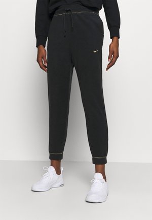 Persoon die zwarte Nike joggers met gouden stiksel en witte sportschoenen draagt tegen een effen achtergrond.