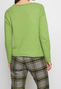 Groen gebreide trui met een brede halslijn, gecombineerd met grijze en gele geruite broek met een subtiel patroon.