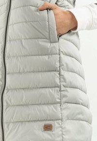 Lichtgrijze gewatteerde bodywarmer met verticale stiknaden, zijzak en een bruin leren merklipje. Gladde textuur en ritssluiting.