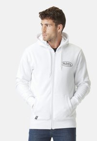 Homme portant un sweat à capuche zippé blanc avec des poches avant et le logo "Von Dutch" sur la poitrine, les mains dans les poches, regardant sur le côté.
