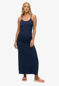 Vestido maxi sin mangas de color azul marino con un corsé ajustado, textura suave y falda larga y fluida. Combinado con sandalias planas rosas con logo.