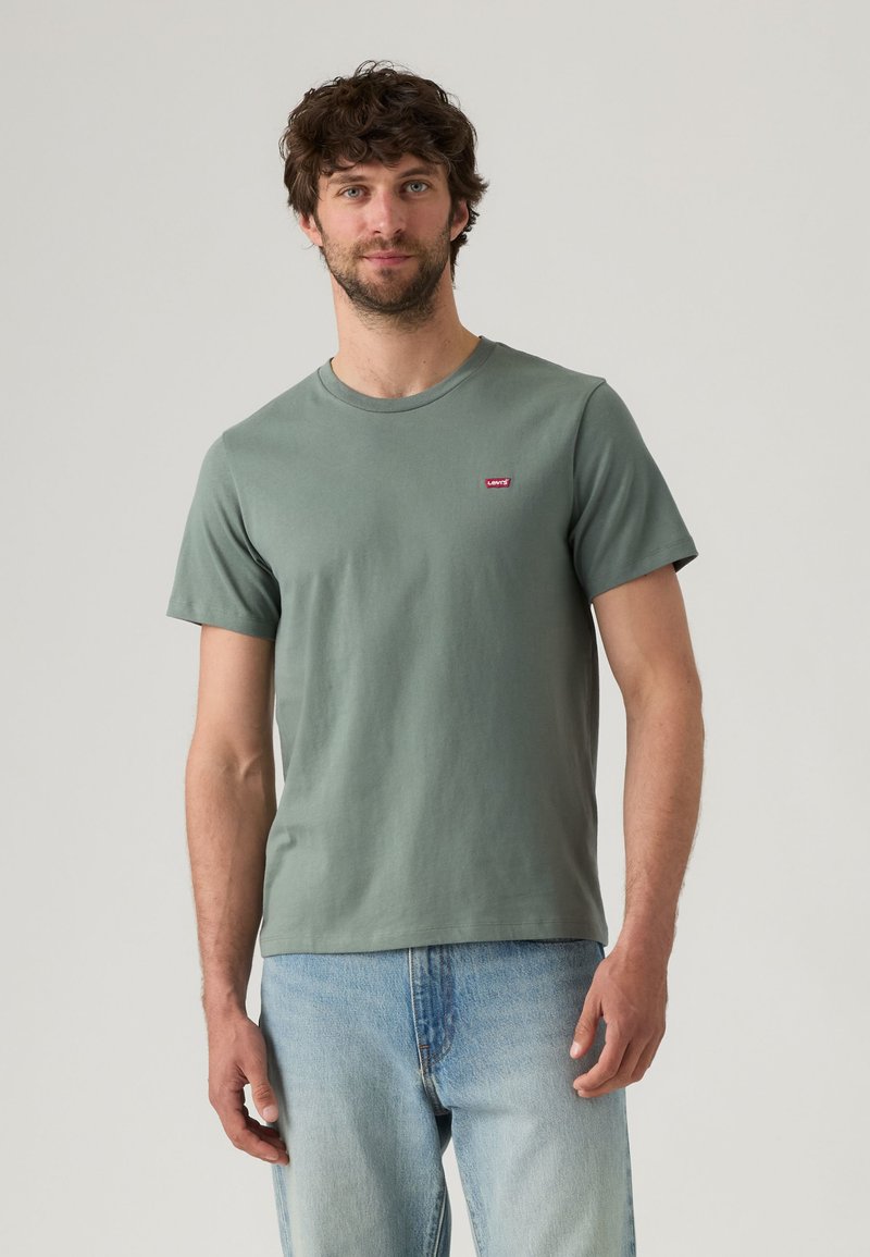 Man with short curly brown hair and beard wears green short-sleeve T-shirt and light blue jeans, standing against plain light background.