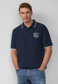 Man in een marineblauwe polo met witte afwerking en de tekst "Service Ford Center", staande met zijn handen in zijn zakken tegen een effen achtergrond.