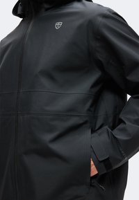 Personne portant une veste noire imperméable avec capuche, fermeture éclair, poches latérales et poignets réglables en velcro.