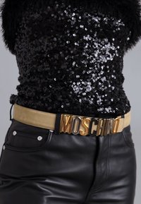 Μπεζ ζώνη με αγκράφα "MOSCHINO" σε χρυσό και μαύρο, συνδυασμένη με μαύρη τοπ με παγιέτες και μαύρο δερμάτινο παντελόνι.
