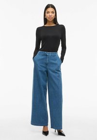 Crni dugi rukav majica u kombinaciji sa visokim širokim plavim trapericama. Crne špicaste štikle upotpunjuju odijelo. Jednostavan, čist dizajn.