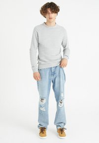 Suéter de canalé gris claro con cuello redondo, combinado con jeans desgastados azul claro que presentan grandes desgarrones. Zapatos marrones de cordones.