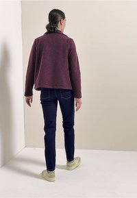 Burgundská texturovaná bunda s límcem, spárovaná s tmavě modrými džíny a světlými zelenými teniskami. Oblečení se vyznačuje jednoduchými, čistými liniemi.