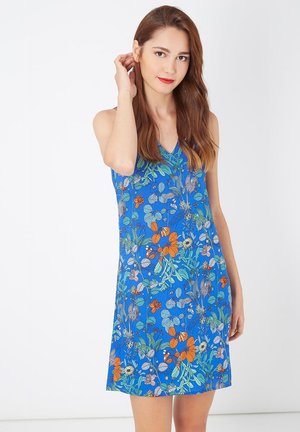Joven mujer con cabello largo y marrón, vistiendo un vestido sin mangas de color azul con un patrón floral colorido, de pie contra un fondo blanco liso.