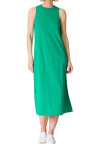Femme portant une robe midi verte sans manches associée à des baskets blanches, debout sur fond blanc.
