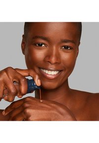A dark-skinned woman applies a liquid product from a sleek blue bottle onto her arm, showcasing smooth skin and even texture.