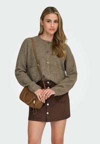 Cardigan en tricot marron avec de grands boutons, associé à une jupe marron avec des poches avant et des boutons-pression. Petit sac à main marron présenté.