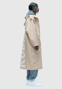 Beżowy wodoodporny trencz z szarym kapturem, ozdobiony dużym białym napisem "KUBAX" oraz zestawiony z jasnoniebieskimi spodniami dresowymi i białymi sneakersami.