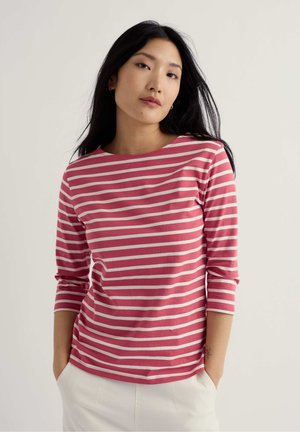 Giovane donna con lunghi capelli neri che indossa una maglietta a righe rosa e bianche con maniche a tre quarti e pantaloni bianchi, con le mani nelle tasche.