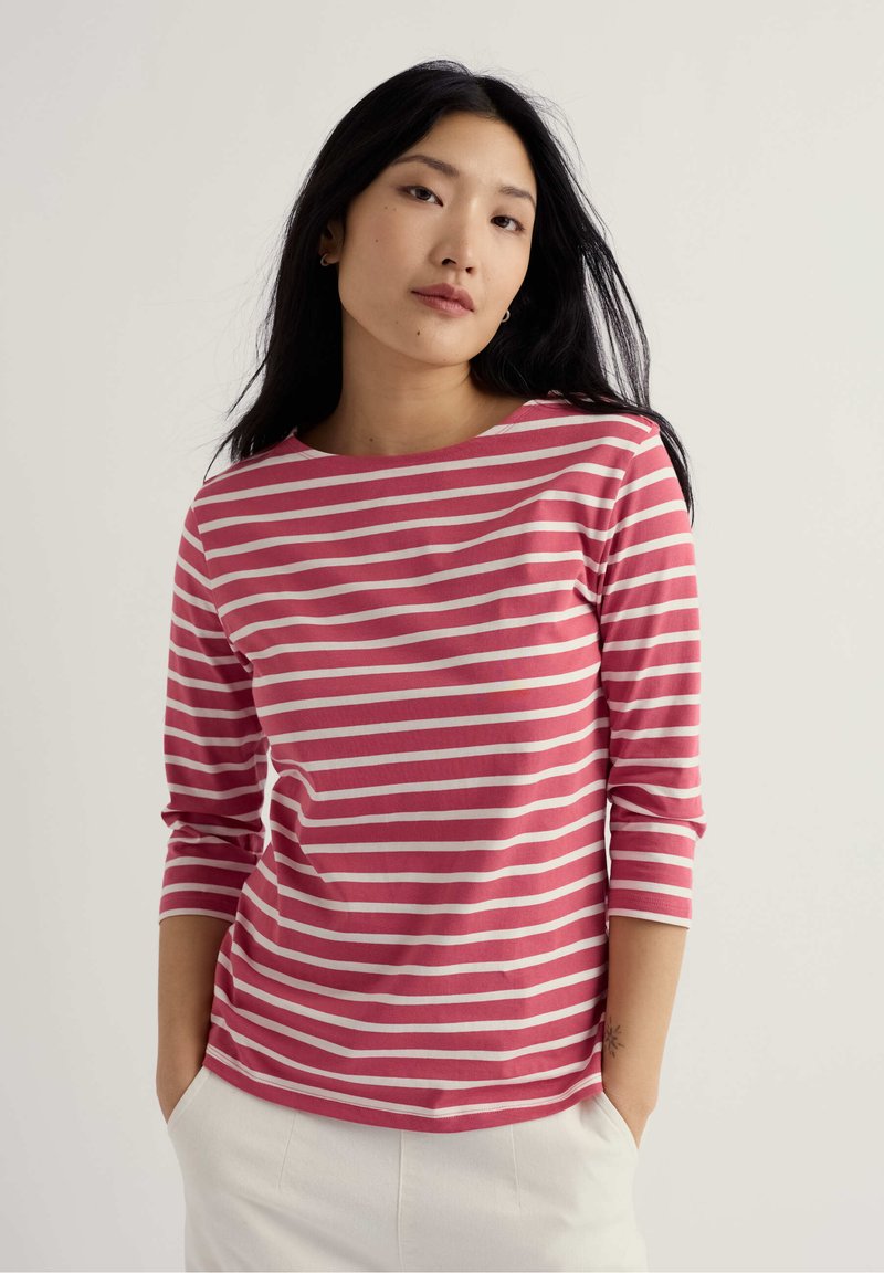 Noor naine, kellel on pikad mustad juuksed, kannab roosa-valgete triipudega kolmeveerandvarrukatega särki ja valgeid pükse, käed taskutes.