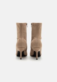 Bottes ankle en daim beige avec un talon haut fin. Présente une fermeture éclair à l'arrière et des coutures visibles à l'extérieur.