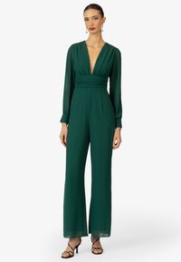 Groene jumpsuit met lange mouwen, een diepe V-hals en een geraffineerde taille. Heeft een wijde pijpen, zachte stof en doorschijnende mouwmanchetten. Zwarte strappy hakken.